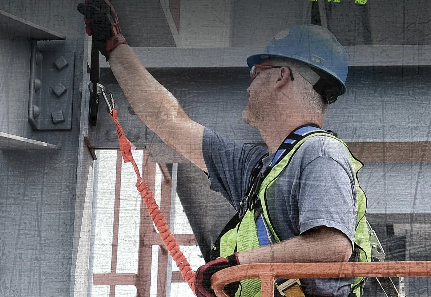 Ironworker on a scaffold reaching overhead, his tool tethered back to his harness by a lanyard